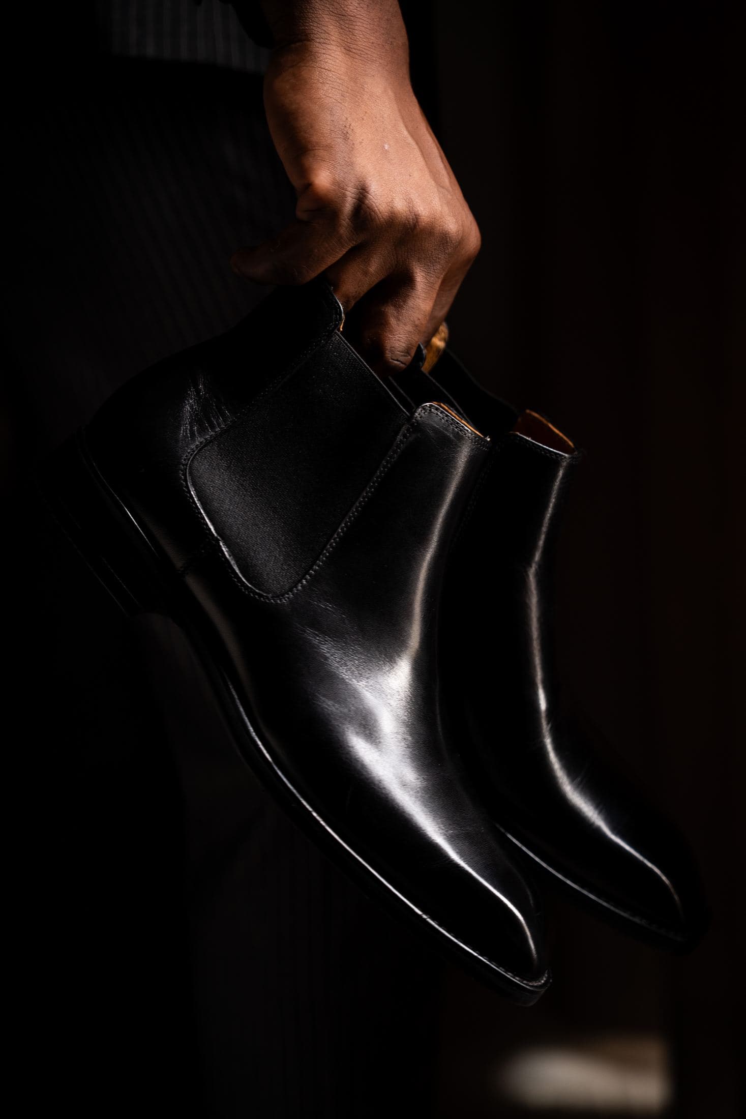 The same hand holding a pair of black Chelsea boots against the dark